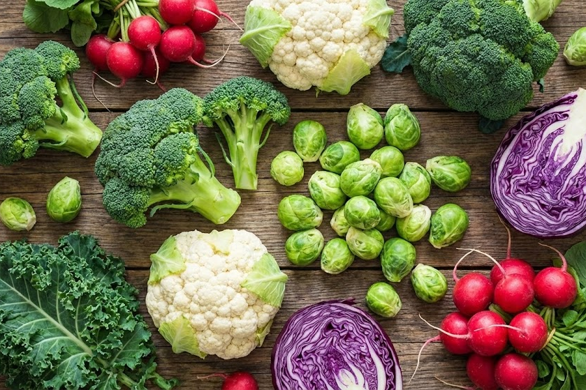Vista superior de uma variedade de vegetais crucíferos frescos dispostos sobre uma mesa de madeira rústica, incluindo brócolis, couve-flor branca e roxa, couve-de-bruxelas, couve folha, repolho roxo e rabanetes