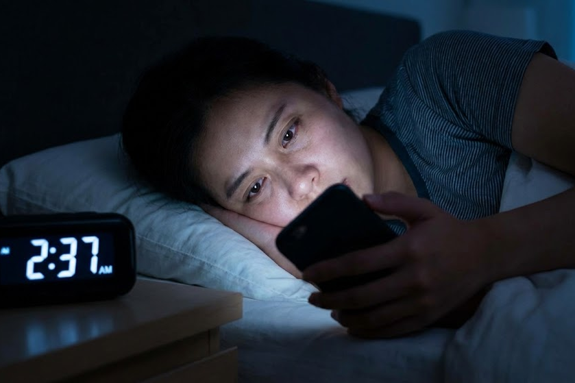 Close-up de uma mulher deitada na cama em um quarto escuro às 2:37 da manhã, com o rosto iluminado pela luz do smartphone que ela está usando, com olhos cansados, representando a procrastinação vingativa do sono e o vício em tecnologia.