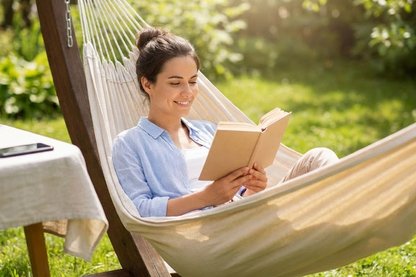 Uma mulher sorrindo e relaxada, deitada em uma rede em um jardim ensolarado, lendo um livro físico de papel. Um smartphone está visível em uma mesa distante, virado para baixo, simbolizando um detox digital bem-sucedido e bem-estar mental.